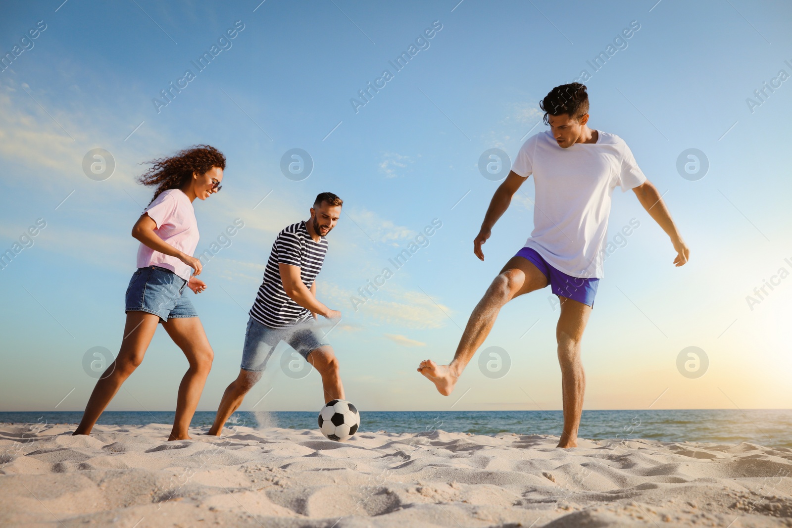 Image of Happy friends playing football on beach during sunset, low angle view