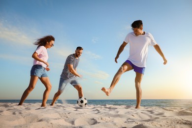 Image of Happy friends playing football on beach during sunset, low angle view