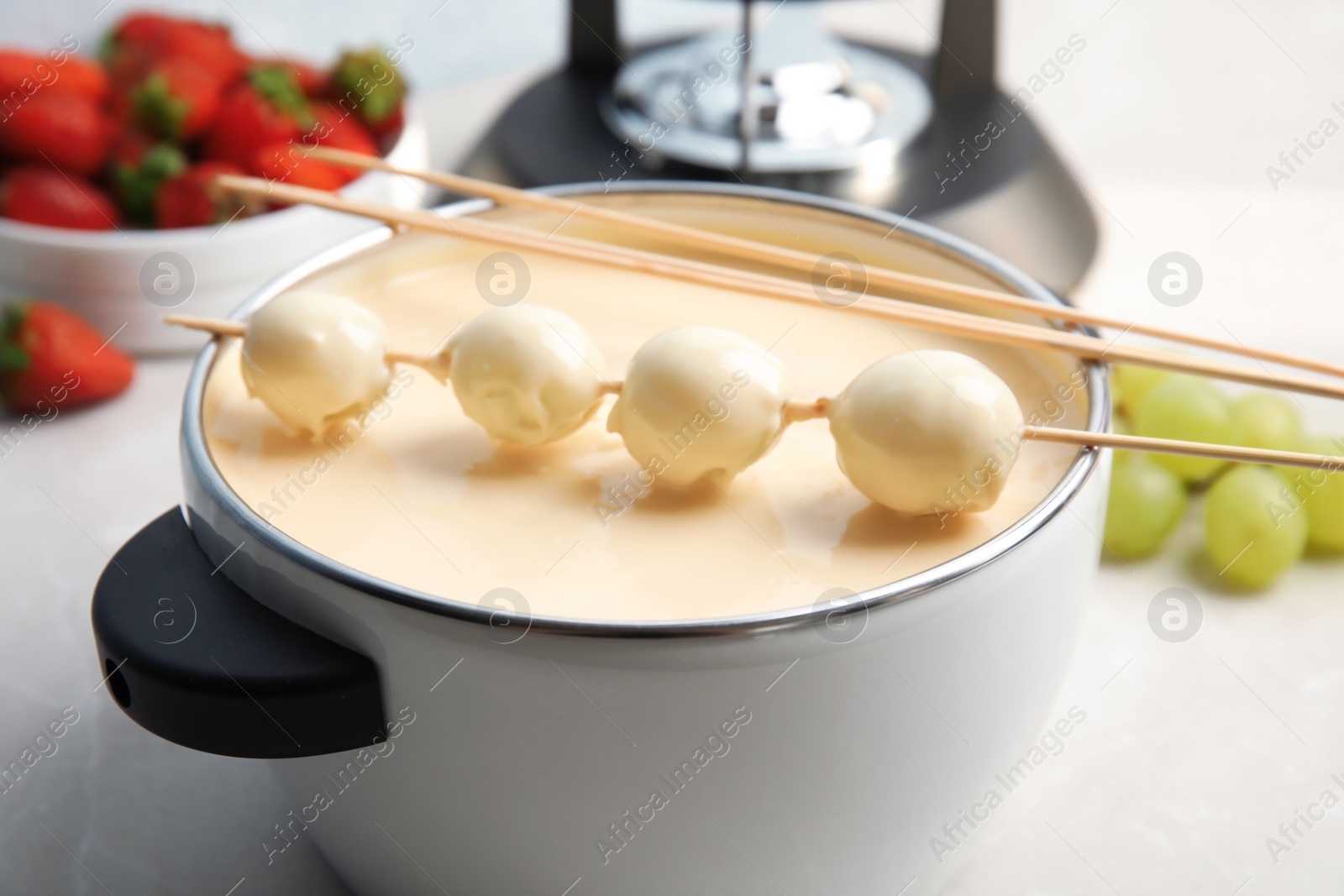 Photo of Pot with white chocolate fondue, dipped grapes and sticks on table, closeup