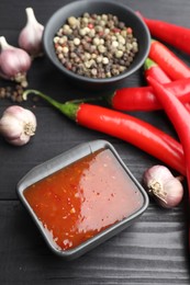 Photo of Spicy chili sauce, garlic, peppers and peppercorns on black wooden table