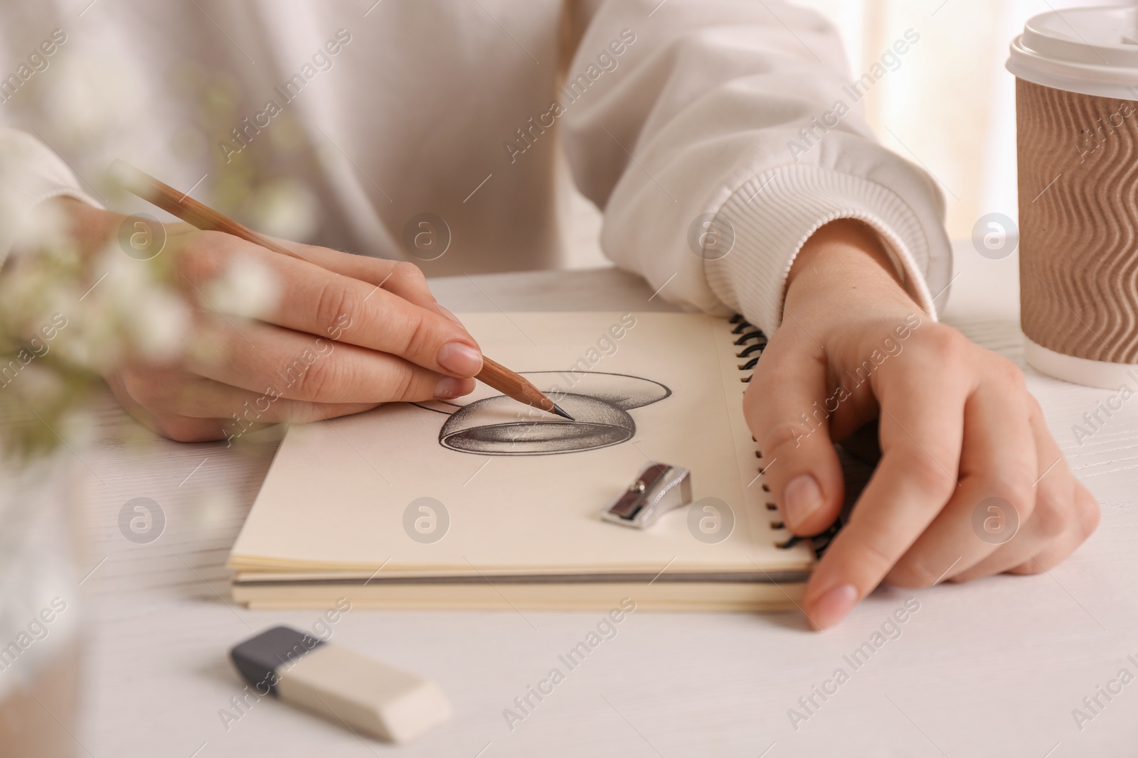Photo of Woman drawing cup of drink with graphite pencil in sketchbook at table, closeup