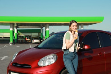 Photo of Beautiful young woman with coffee eating hot dog near car at gas station