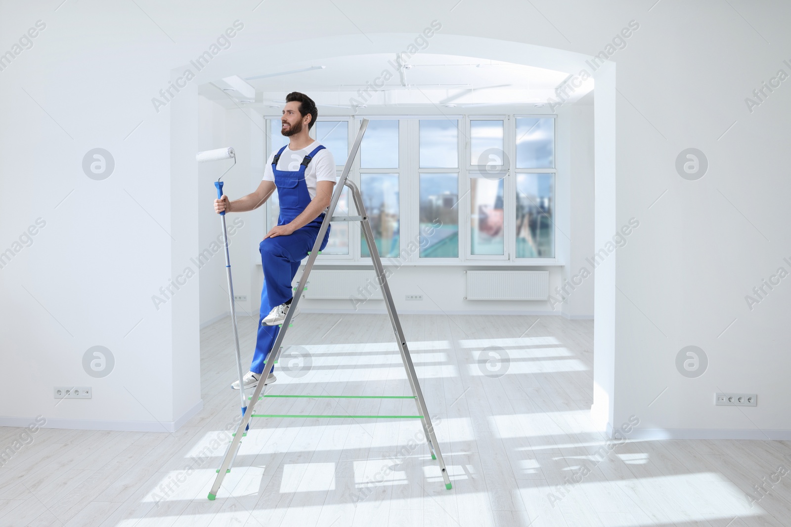 Photo of Handyman with roller on step ladder in room. Ceiling painting