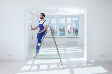 Photo of Handyman with roller on step ladder in room. Ceiling painting