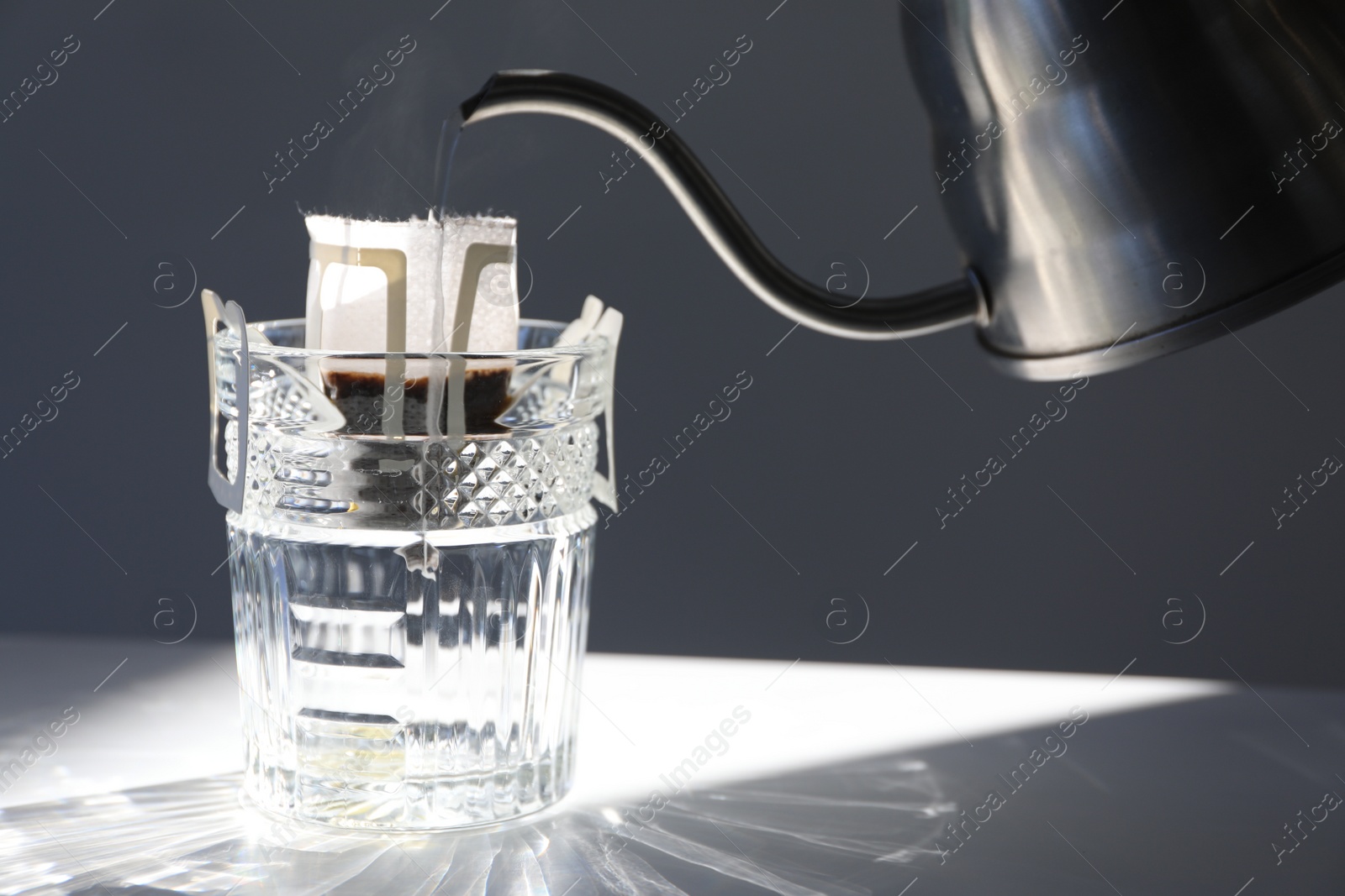 Photo of Pouring hot water into glass with drip coffee bag from kettle on light grey table, closeup