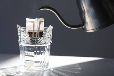 Photo of Pouring hot water into glass with drip coffee bag from kettle on light grey table, closeup