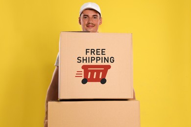 Image of Happy young courier with cardboard boxes on yellow background. Free shipping