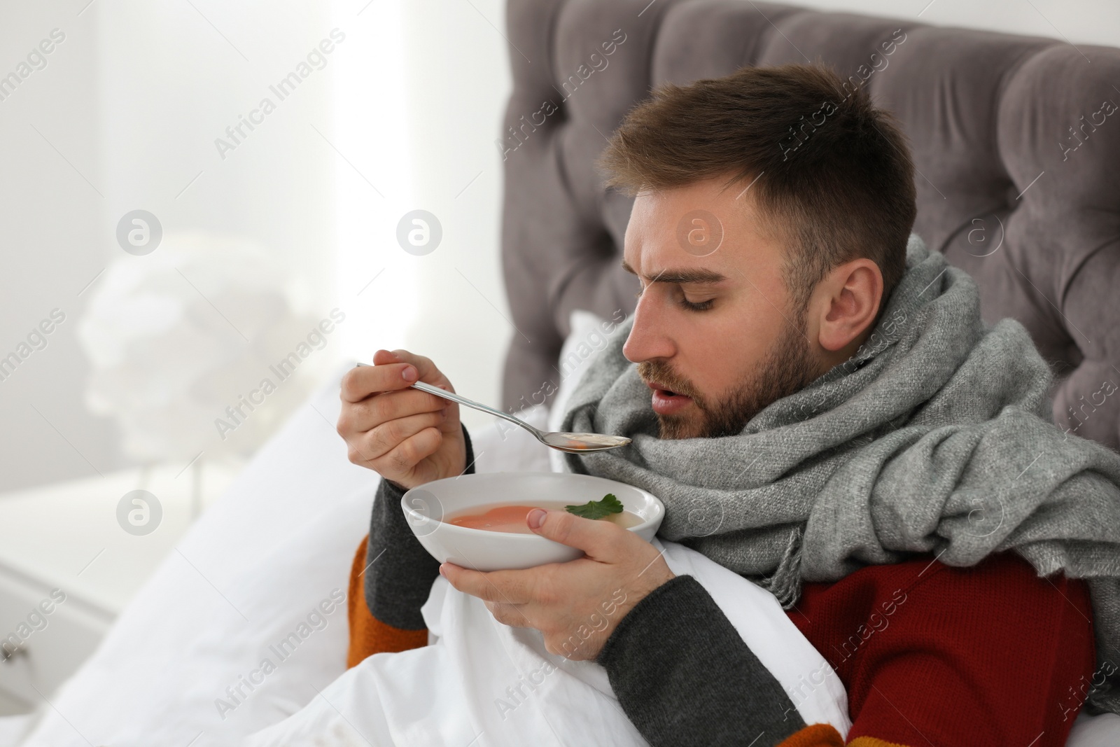 Photo of Sick young man with bowl of tasty soup in bed at home