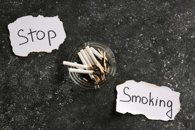 Photo of Ashtray with burnt cigarettes and words Stop Smoking written on paper on black textured table, flat lay