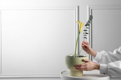 Photo of Stylish ikebana as house decor. Woman creating floral composition with calla flower and eucalyptus branch at table near white wall, closeup and space for text