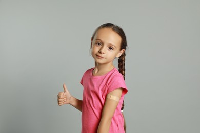 Photo of Vaccinated little girl with medical plaster on her arm showing thumb up against light grey background