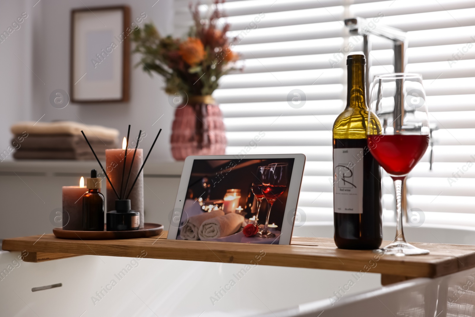 Photo of Wooden tray with tablet, wine and burning candles on bathtub in bathroom