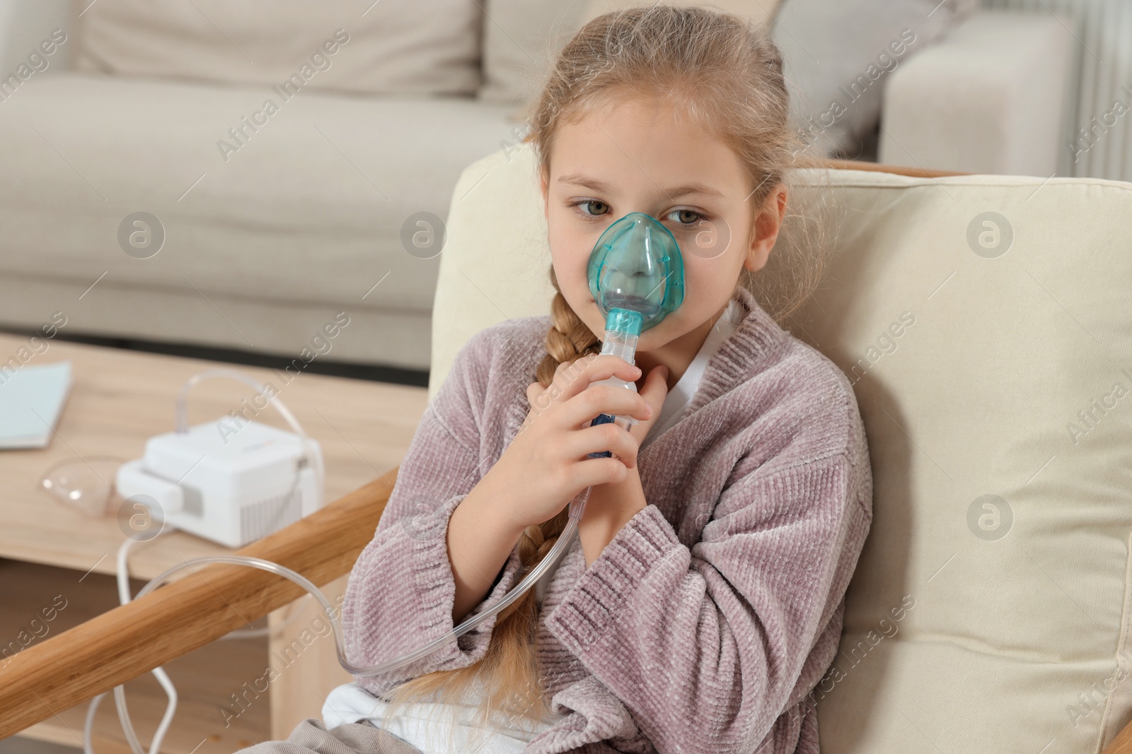 Photo of Little girl using nebulizer for inhalation in armchair at home