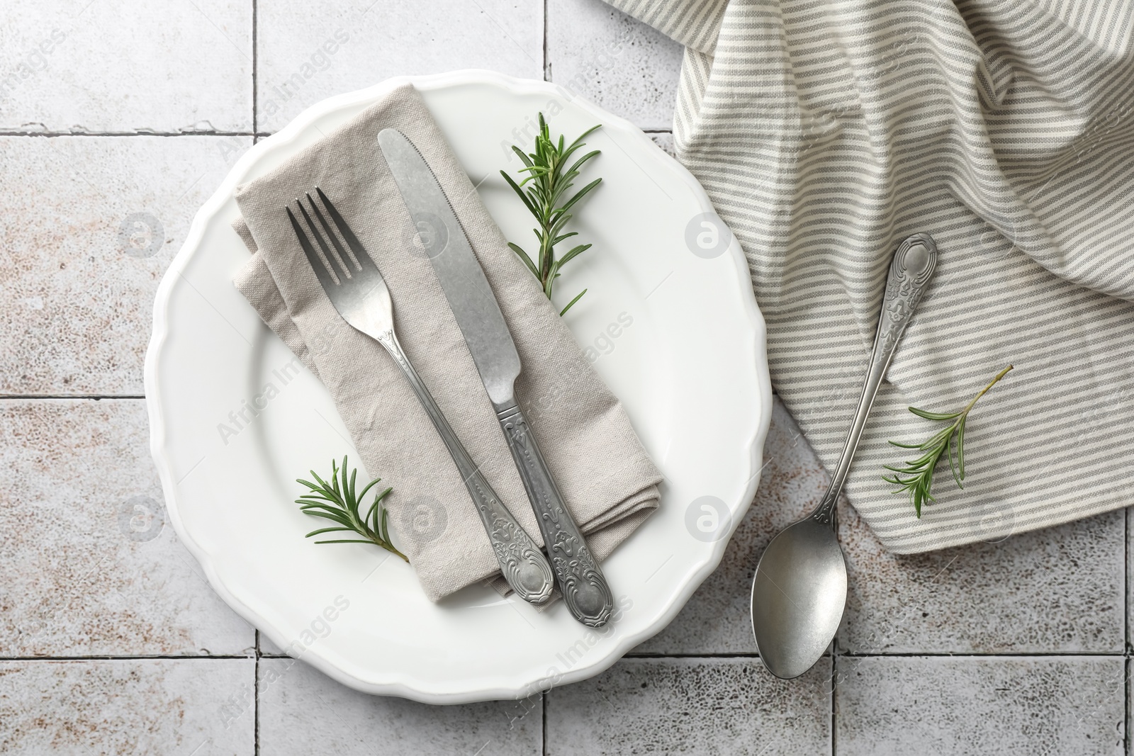 Photo of Stylish setting with cutlery, napkin, rosemary and plate on light tiled table, top view