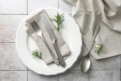 Stylish setting with cutlery, napkin, rosemary and plate on light tiled table, top view