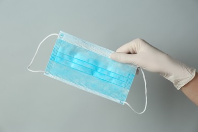 Doctor in medical gloves holding protective mask on light grey background, closeup