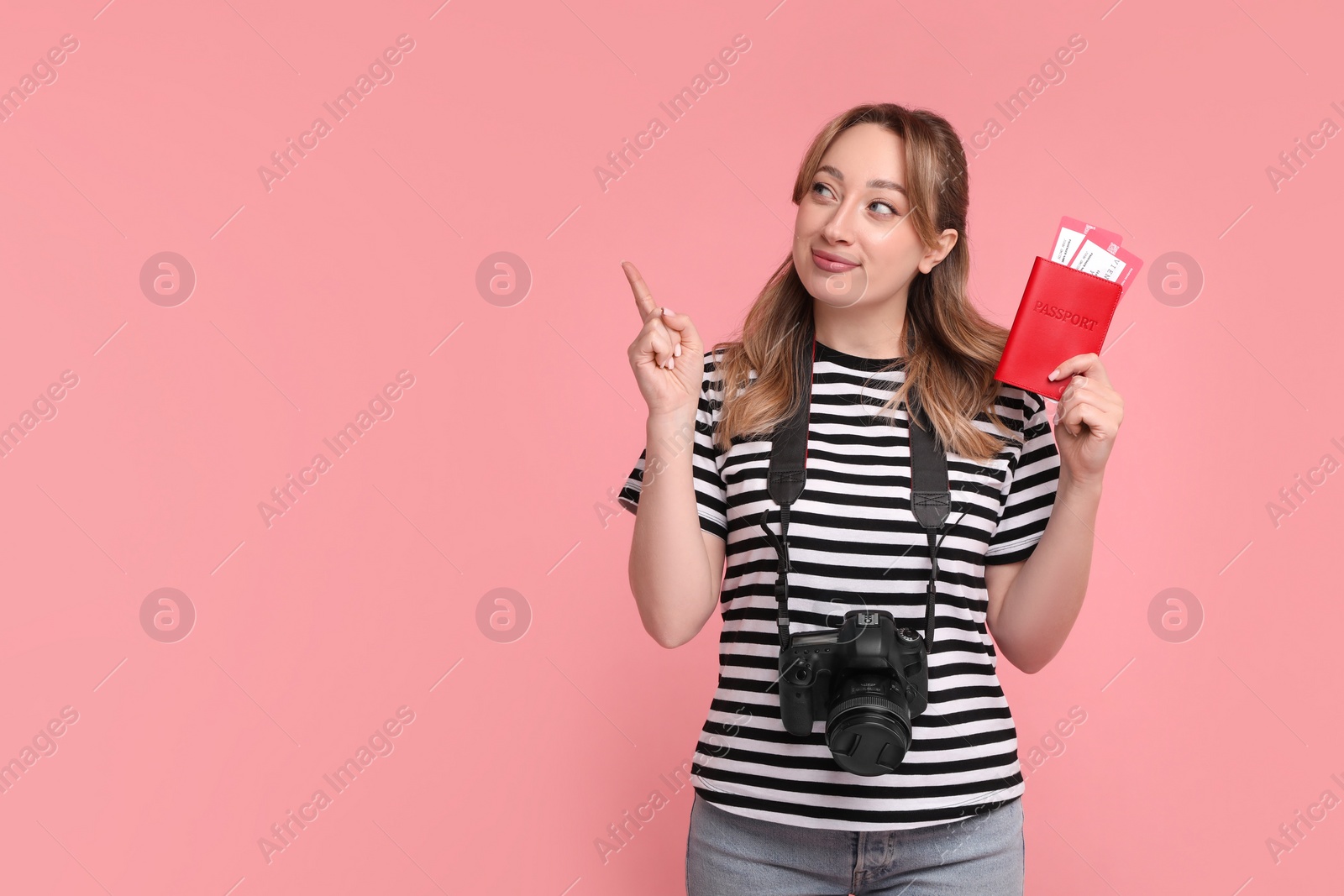 Photo of Happy young woman with passport, ticket and camera pointing at something on pink background, space for text