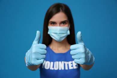 Photo of Female volunteer in protective mask and gloves showing thumbs up gesture against blue background, focus on hands. Aid during coronavirus quarantine	