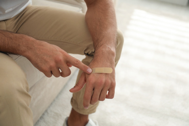 Photo of Man putting sticking plaster onto hand indoors, closeup