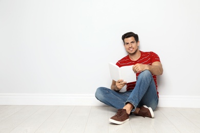 Handsome man reading book on floor near white wall, space for text