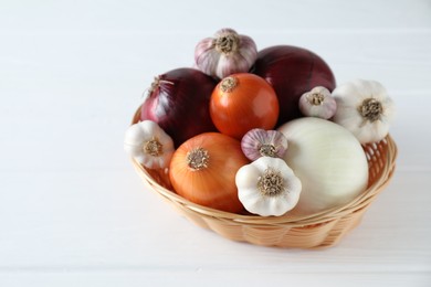 Fresh raw garlic and onions in wicker basket on white table, closeup. Space for text