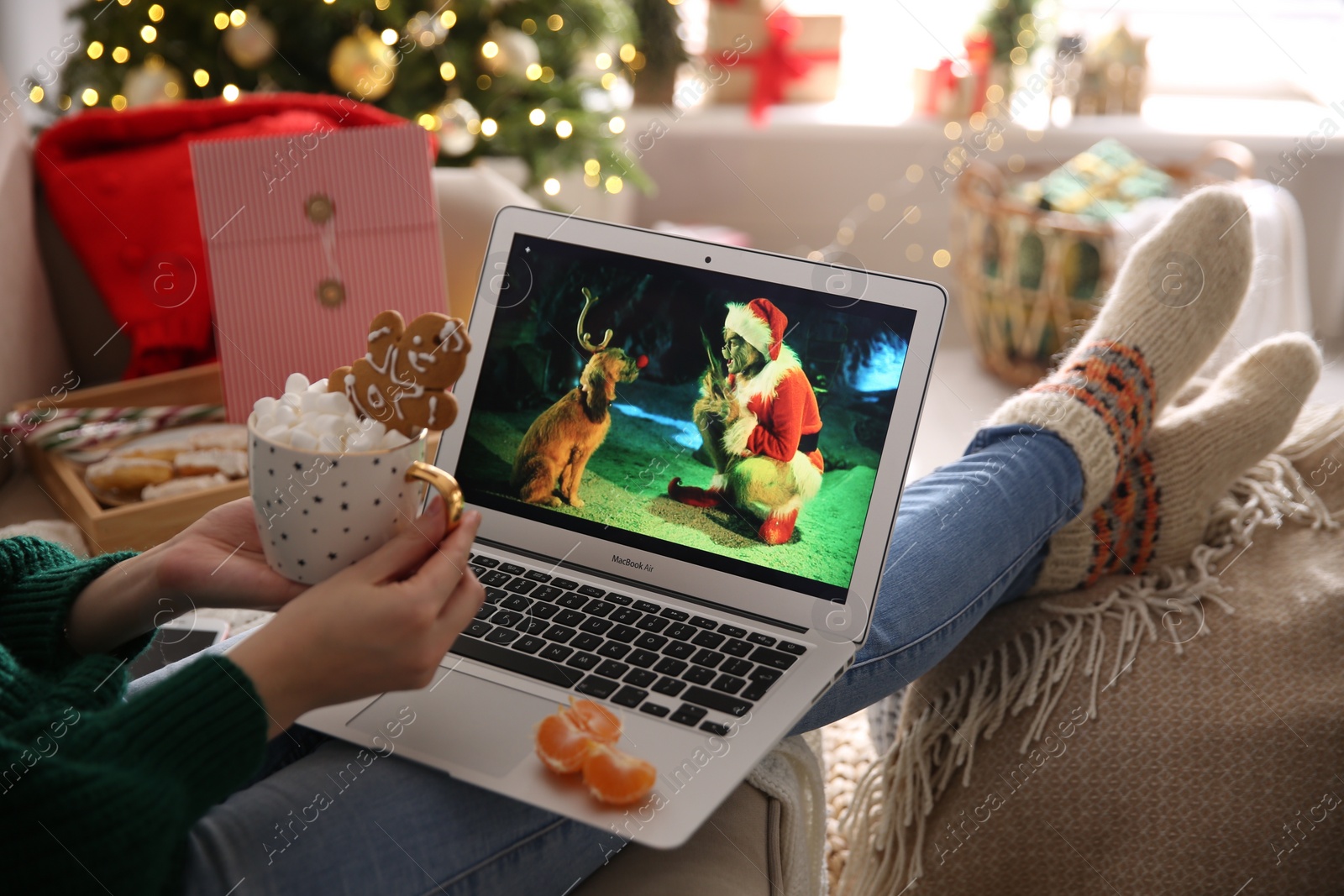 Photo of MYKOLAIV, UKRAINE - DECEMBER 25, 2020: Woman with sweet drink watching The Grinch movie on laptop at home, closeup. Cozy winter holidays atmosphere