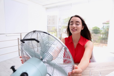 Photo of Young woman enjoying air flow from fan at workplace. Summer heat