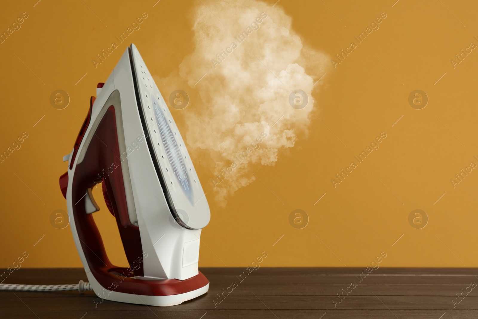 Image of Modern iron with steam on wooden table against orange background, space for text