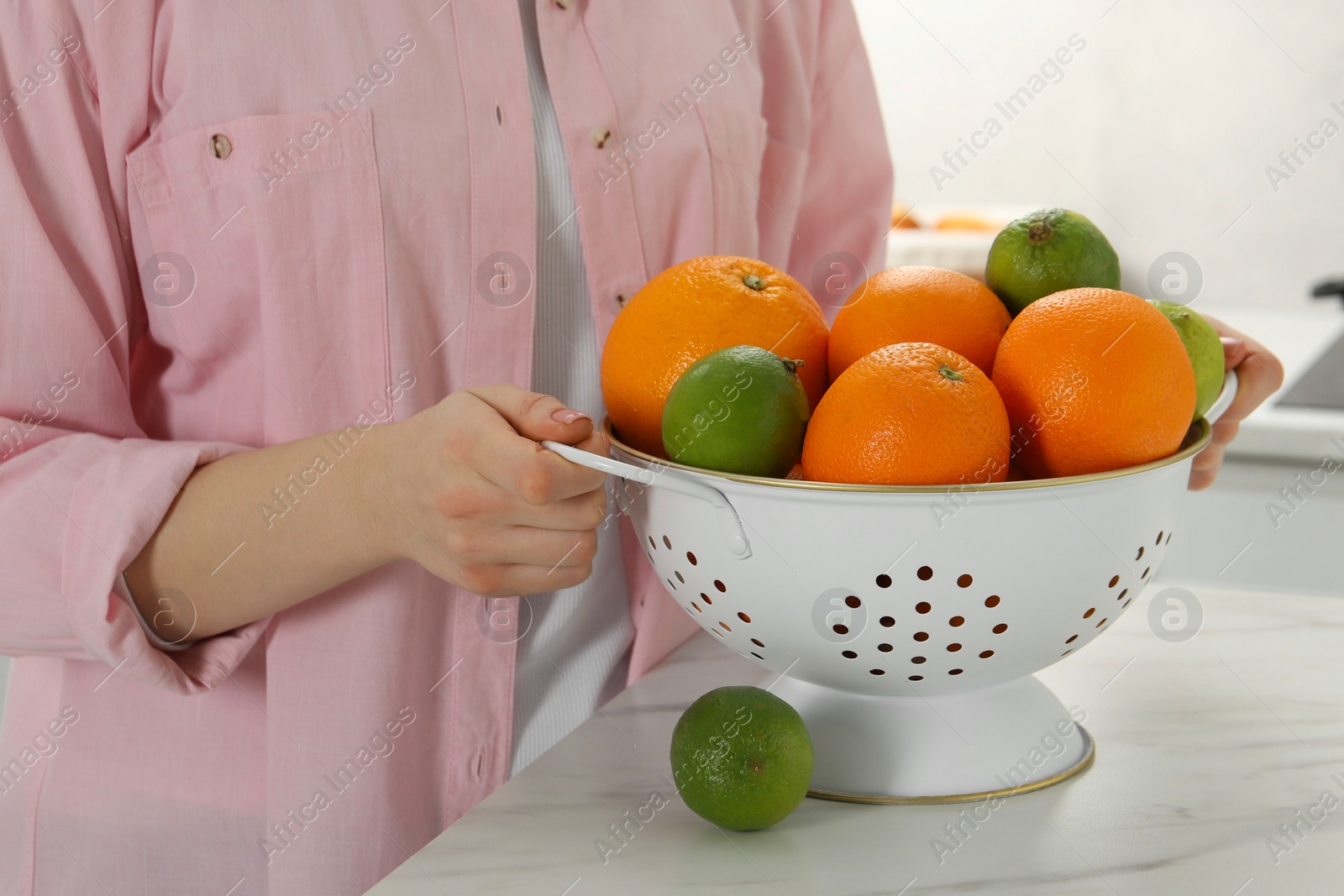 Photo of Woman holding colander with fresh fruits at white marble table in kitchen, closeup