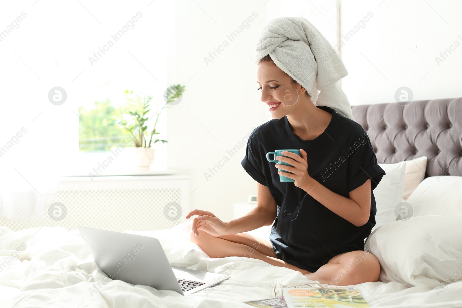 Photo of Beautiful young woman with cup of coffee and laptop sitting in bed at home. Lazy morning