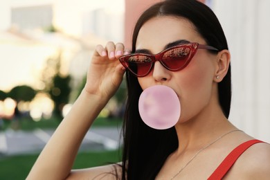 Photo of Beautiful woman in stylish sunglasses blowing gum near building outdoors