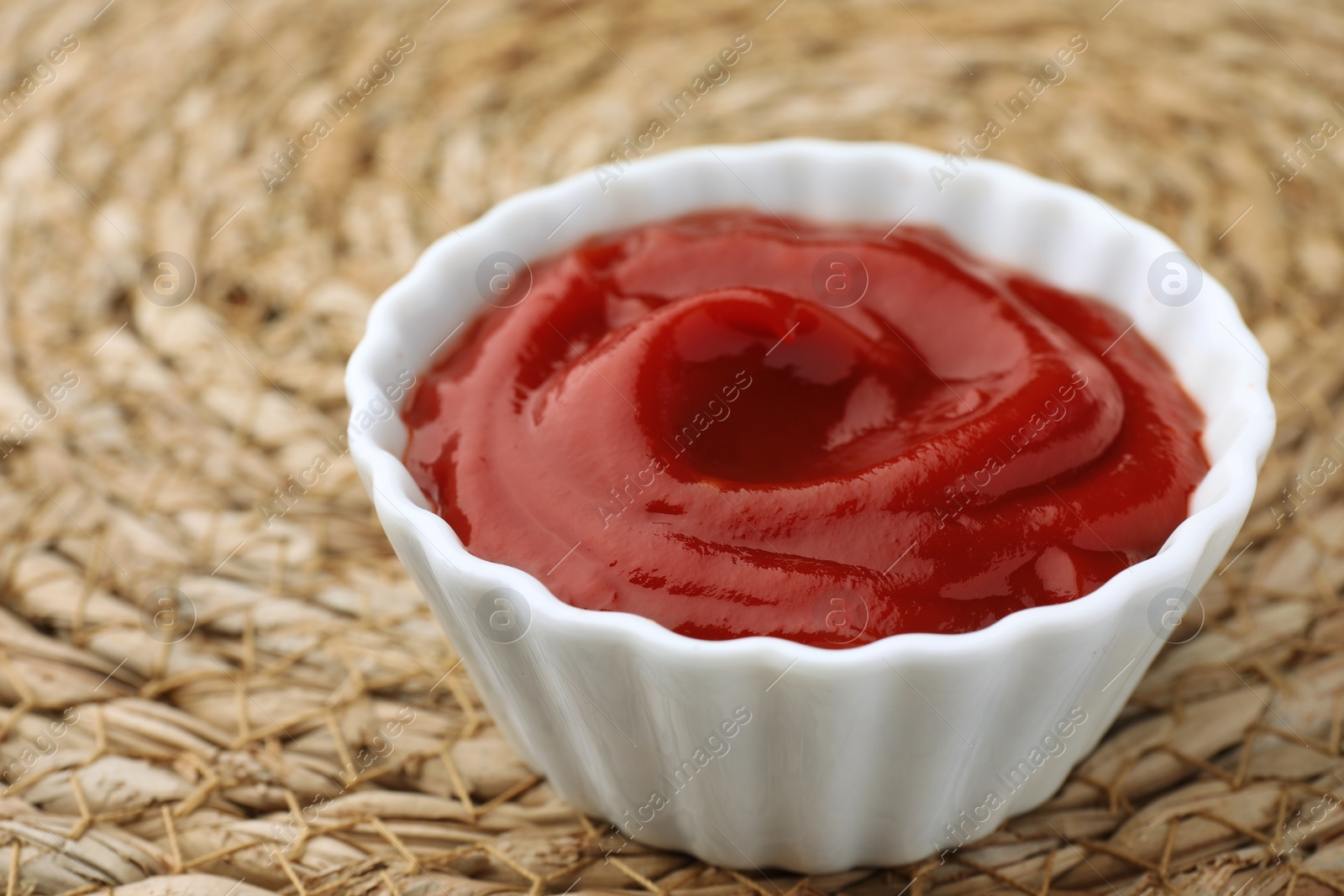 Photo of Bowl of tasty ketchup on wicker mat, closeup