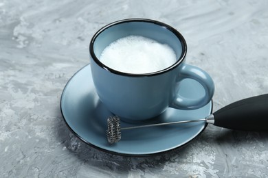 Photo of Mini mixer (milk frother) and whipped milk in cup on grey textured table, closeup