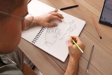 Man drawing in sketchbook with pencil at wooden table, closeup