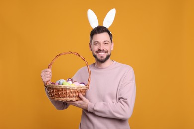 Photo of Happy man in bunny ears headband holding wicker basket with painted Easter eggs on orange background