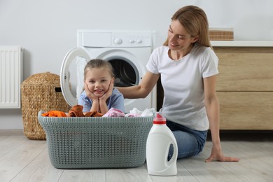 Photo of Mother and daughter sitting near basket with dirty clothes and fabric softener in bathroom