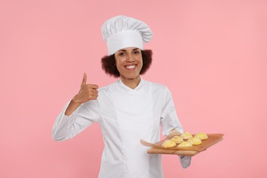 Photo of Happy female chef in uniform holding board with cookies and showing thumb up on pink background