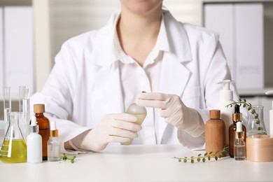 Photo of Dermatologist with bottle of cosmetic product at white table indoors, closeup. Development and testing