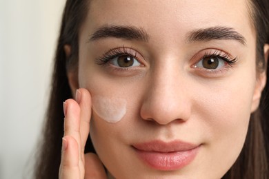 Young woman with dry skin applying cream onto her face on blurred background, closeup