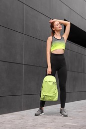 Photo of Beautiful woman in gym clothes posing with stylish backpack near dark grey wall on street