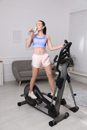 Photo of Happy young woman drinking water during training on elliptical machine at home
