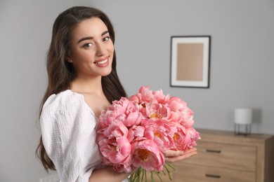 Photo of Beautiful young woman with bouquet of pink peonies at home