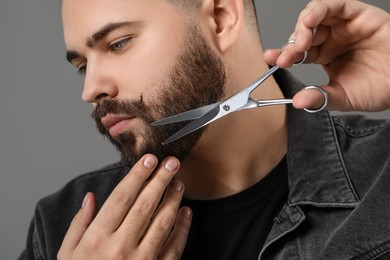 Photo of Handsome young man trimming beard with scissors on grey background, closeup