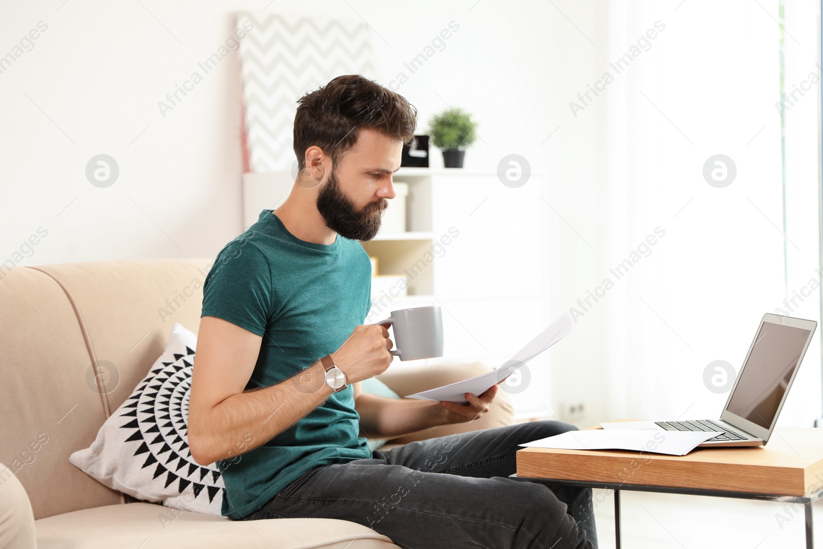 Photo of Young bearded man with coffee and documents working on sofa at home