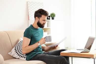 Young bearded man with coffee and documents working on sofa at home