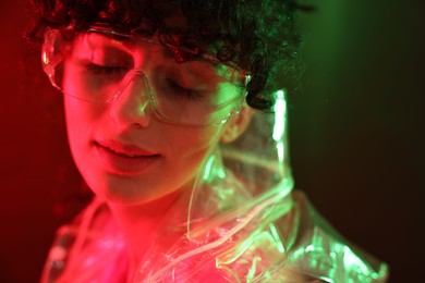 Photo of Beautiful young woman in transparent coat and sunglasses posing on color background in neon lights, closeup