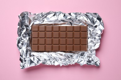 Photo of Delicious milk chocolate bar with foil on light pink background, top view