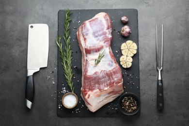 Photo of Piece of raw pork belly, rosemary, garlic, spices, butcher knife and carving fork on grey textured table, flat lay