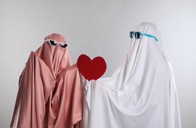 Photo of Cute ghosts. Couple in color sheets and sunglasses with red heart on light grey background