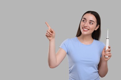 Photo of Happy young woman holding electric toothbrush and pointing on light grey background, space for text